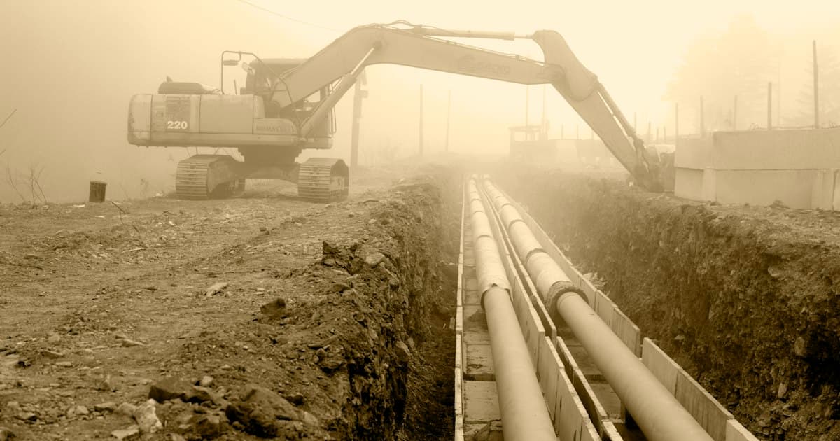 Excavadora junto a una zanja abierta con tuberías de acero, en un día de niebla
